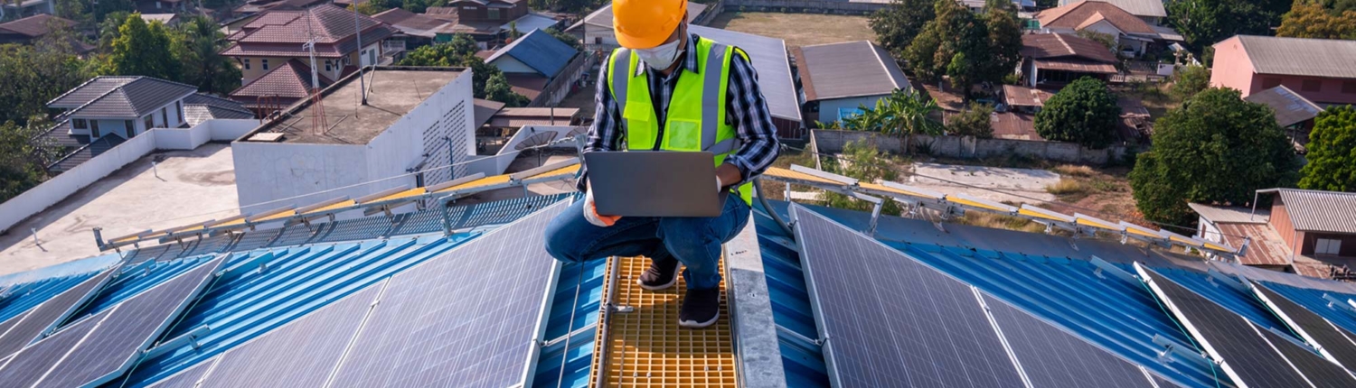 Roofer on top of a roof with solar panels, working on laptop